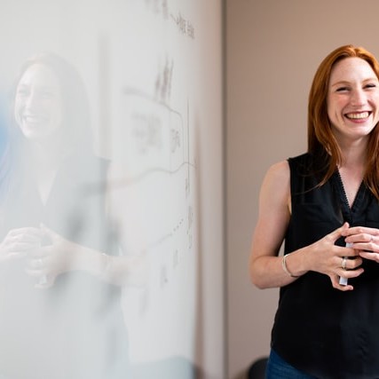 Who we service website image of a white lady with red hair standing by a whiteboard smiling from ear to ear as she grips the marker in both hands.